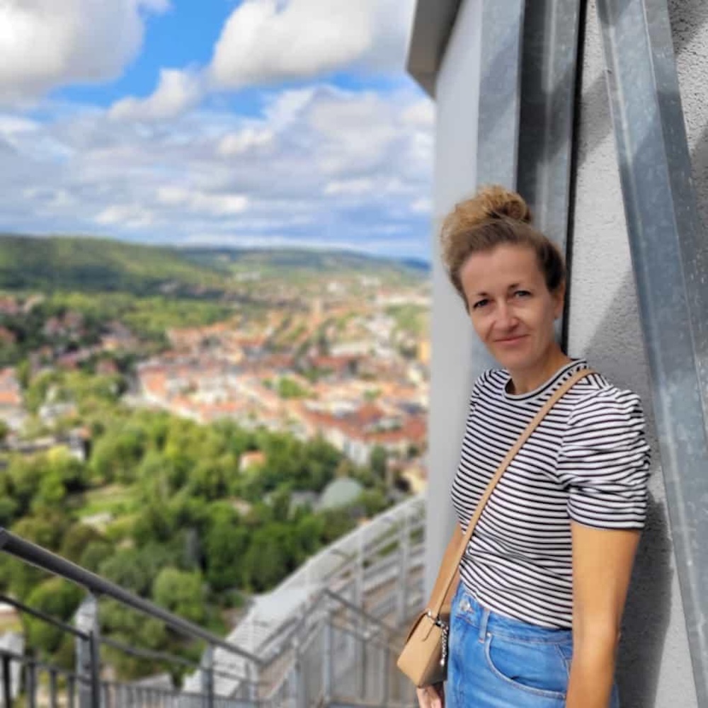 Testimonial und Portrait von Claudia, Teilnehmerin des Online-Programms Mit Klarheit in deine berufliche Zukunft. Sie trägt ein scharz-weißes gestreiftes Shirt, eine Jeans und hat eine hellbraune Umhängetasche. Sie steht auf einer Plattform und Hunter ihr ist ein Stadtpanorama zu sehen.