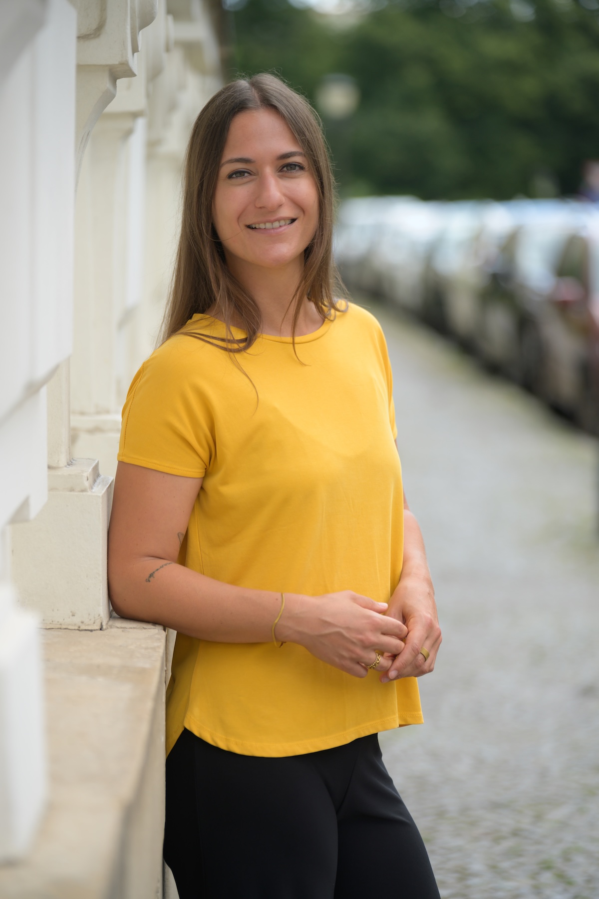 Giulia Barile, DGSv-zertifizierte systemische Supervisorin in Köln und Coach für berufliche Neuorientierung, trägt eingelbes Oberteil und eine schwarze Hose, ist mit dem Ellenbogen an einer Hauswand angelehnt und blickt in die Kamera