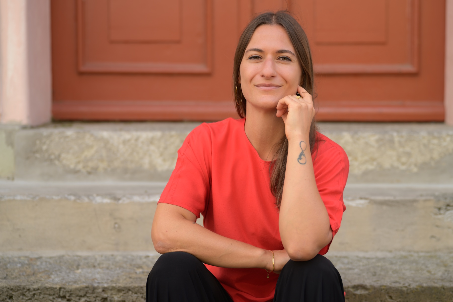 Giulia Barile, systemische Supervisorin in Köln und Coach für berufliche Neuorientierung, sitzt in einem roten Shirt auf einer Treppe vor einem Torbogen