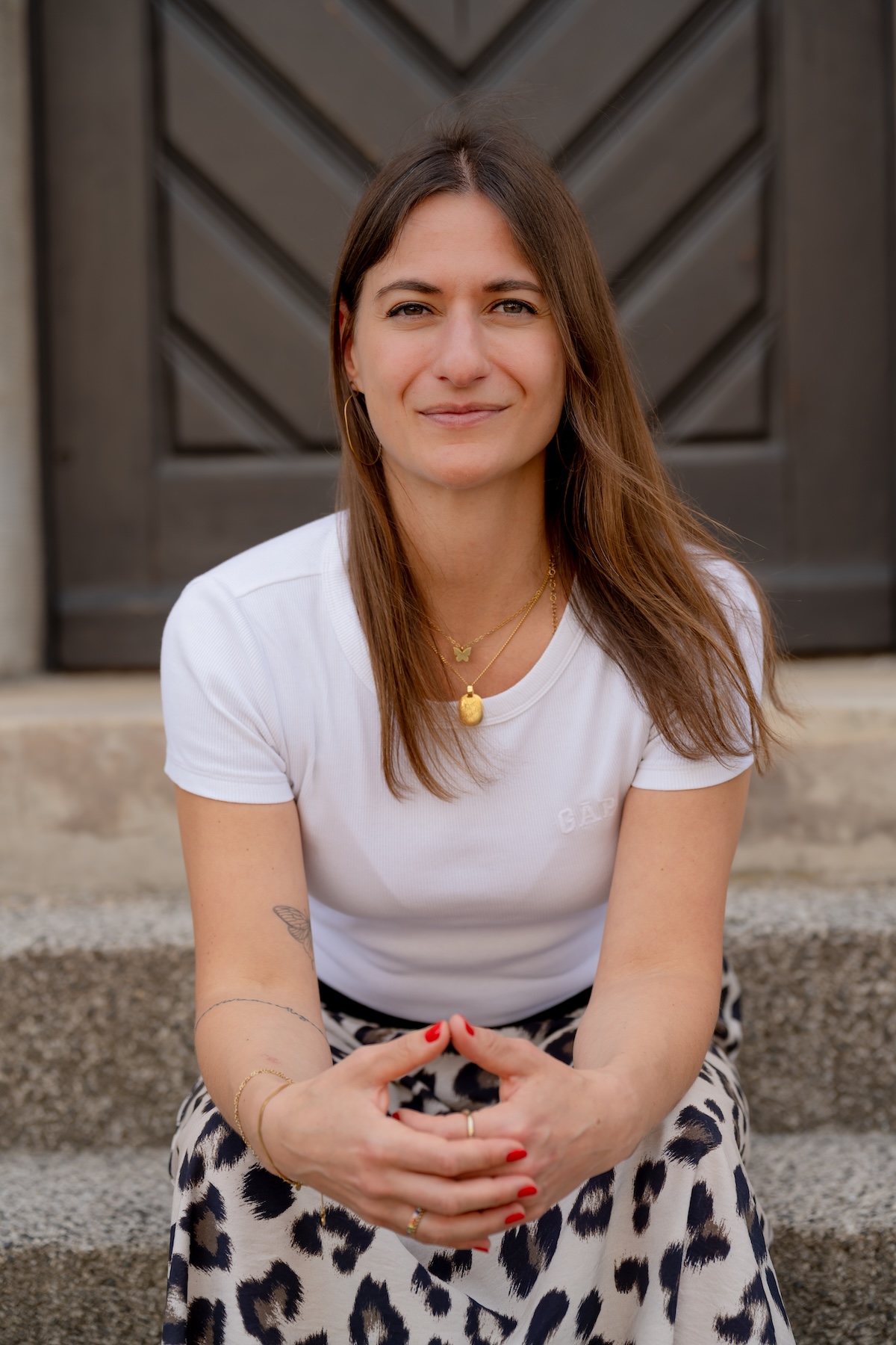 Giulia Barile, DGSv-zertifizierte systemische Supervisorin in Köln und Coach und Mentorin für Frauen in der beruflichen Neuorientierung, trägt ein weißes Shirt, einen Printrock, sitzt auf einer Steintreppe vor einer braunen Holztür