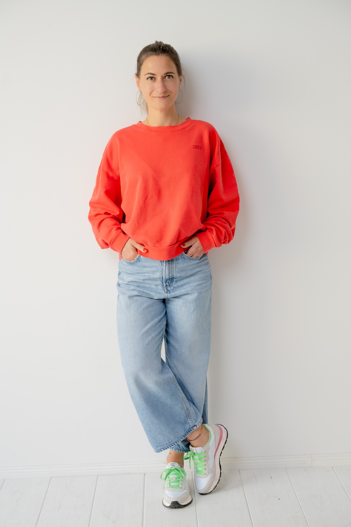 Giulia Barile, DGSv-zertifizierte systemische Supervisorin in Köln und Coach und Mentorin für Frauen in der beruflichen Neuorientierung steht an einer weißen Wand, trägt einen roten Pulli, blaue Jeans und weiße Schuhe mit grünen Schnürsenkeln.