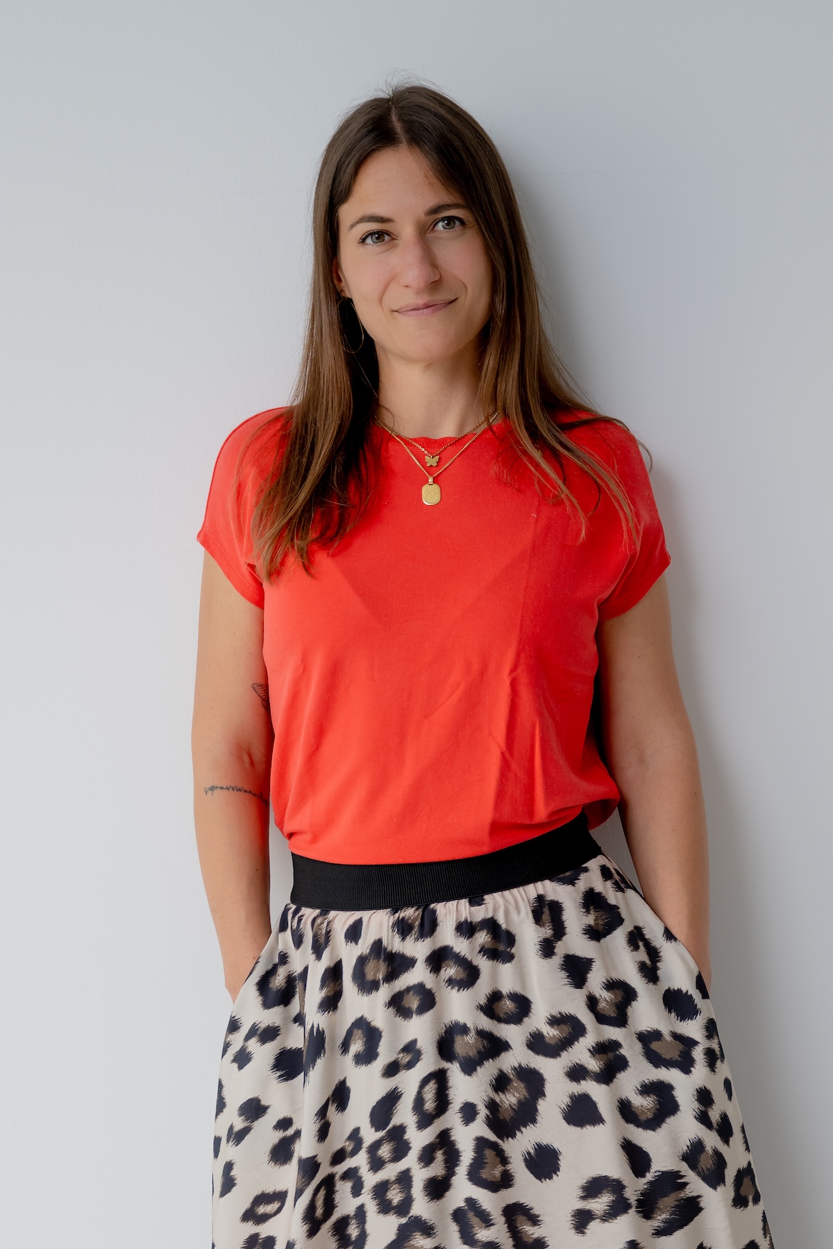 Giulia Barile, DGSv-zertifizierte systemische Supervisorin in Köln und Coach und Mentorin für Frauen in der beruflichen Neuorientierung, trägt ein rotes Shirt, einen knöchellangen Printrock und brauen Sandaletten, stehend an einer weißen Wand