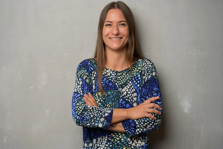 Giulia Barile, DGSv-zertifizierte systemische Supervisorin und Coach, in einem blau, grün, weiß und gelb gesprenkeltem Kleid, kreuzt die Arme voreinander und steht vor einer grauen Wand