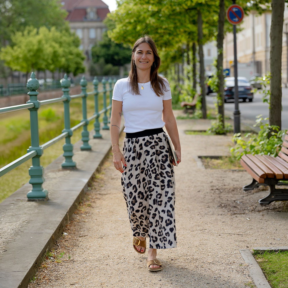 Giulia Barile, DGSv-zertifizierte systemische Supervisorin in Köln und Coach und Mentorin für Frauen in der beruflichen Neuorientierung, trägt ein weißes Shirt, einen knöchellangen Printrock, hat ihr Laptop in der linken Hand und läuft an einem Kanal entlang